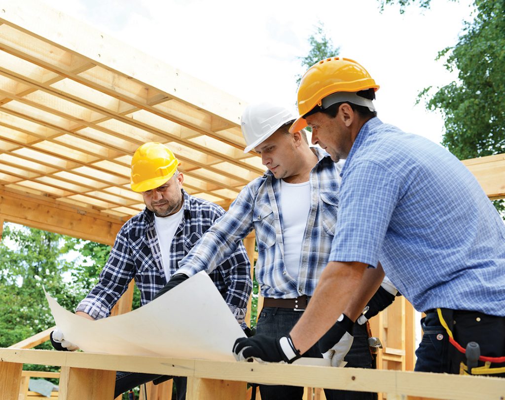 construction workers looking at blueprint at a worksite and wood framed house.
